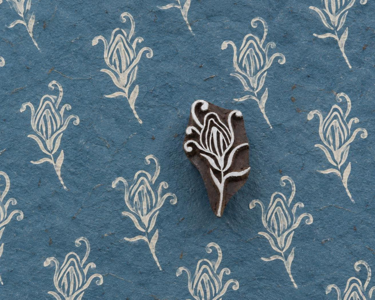 Hand carved Indian wooden printing block with a curling tulip motif beside repeated white impressions on textured blue handmade paper
