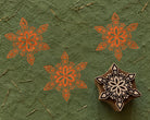 Folk Snowflake hand carved wooden printing block with repeated orange snowflake impressions on green handmade paper.