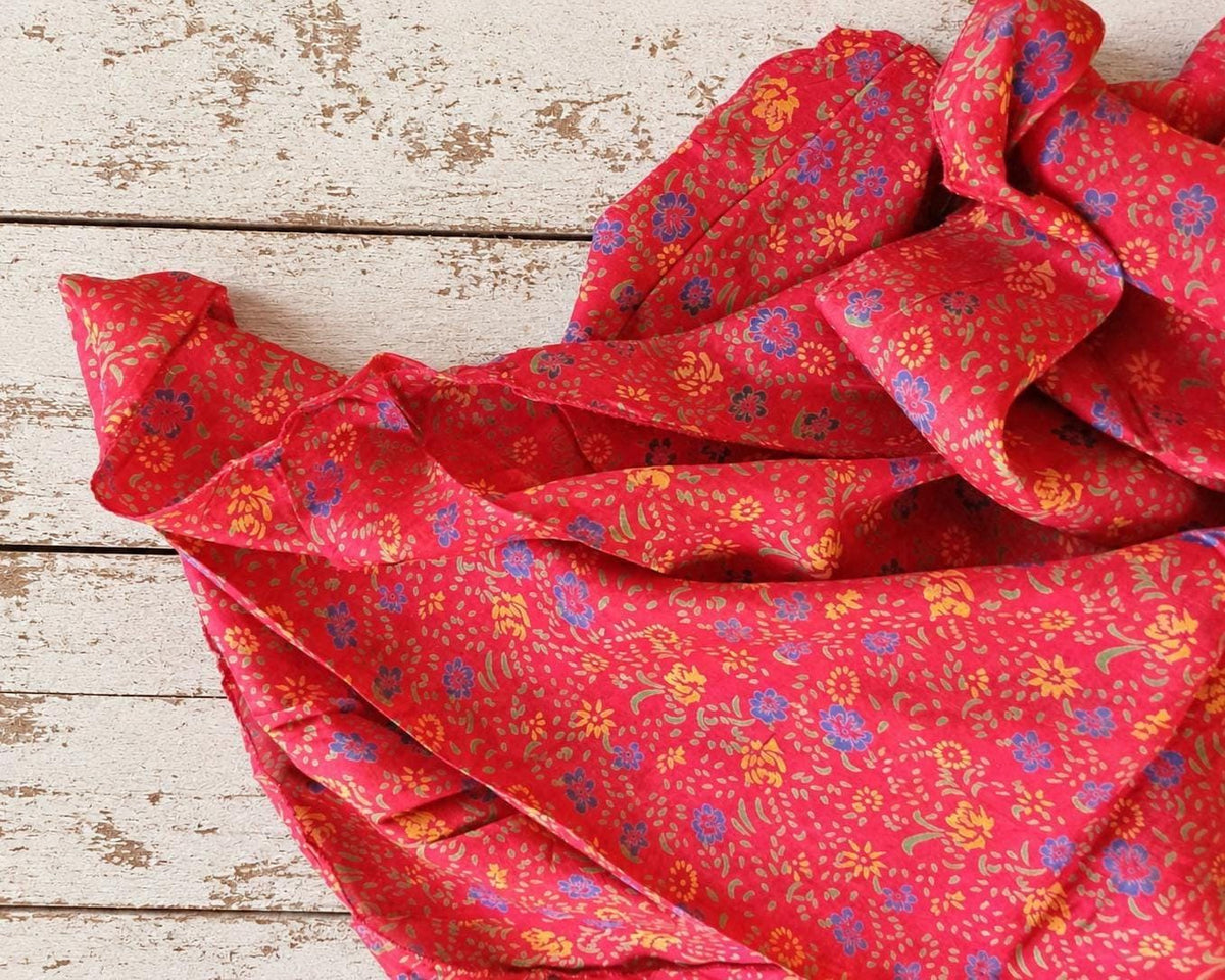 Vintage coral-red sari silk fabric with small blue and yellow flowers, folded on a rustic white wooden surface.