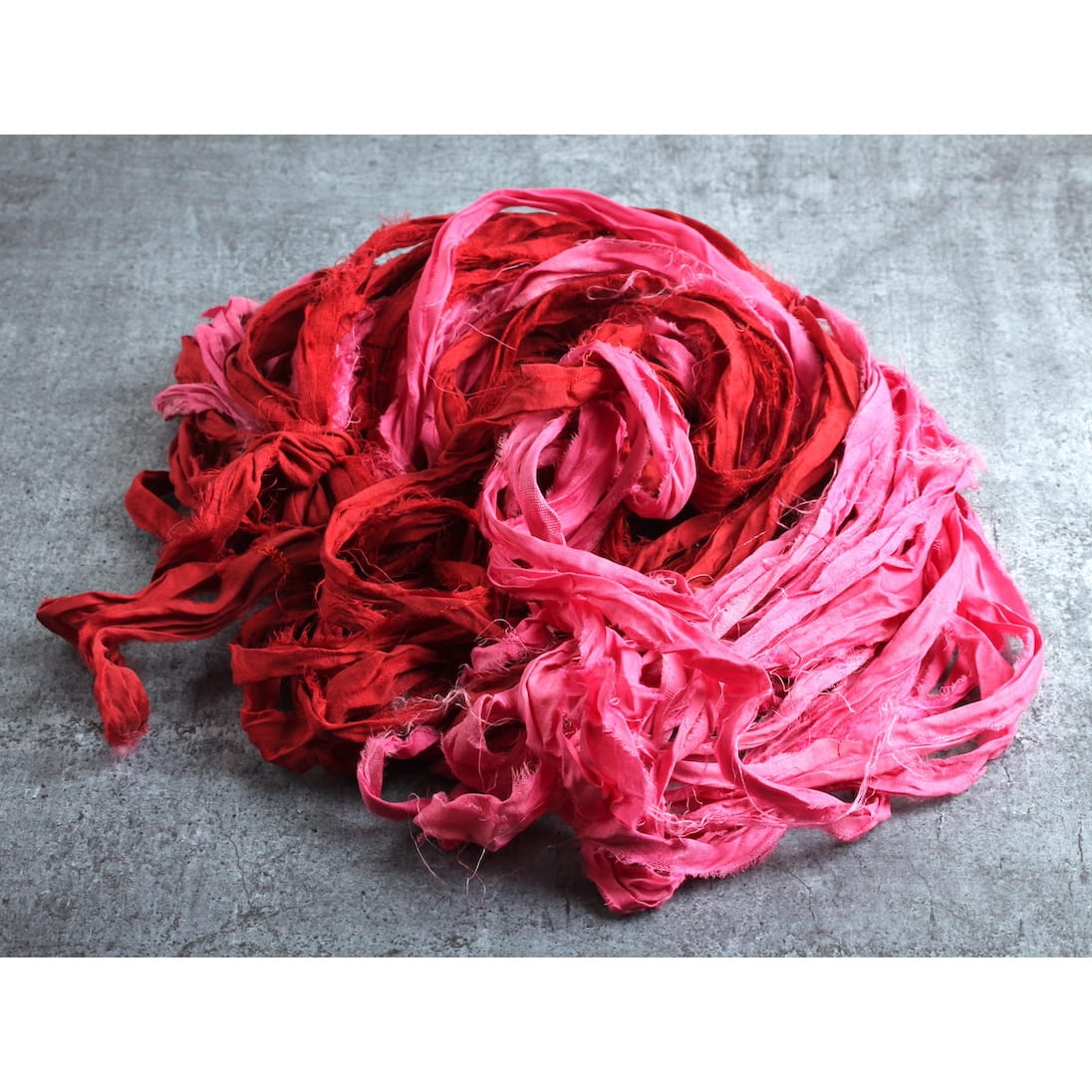 Red and pink sari silk ribbon displayed open, showing handmade frayed texture and recycled silk charm.